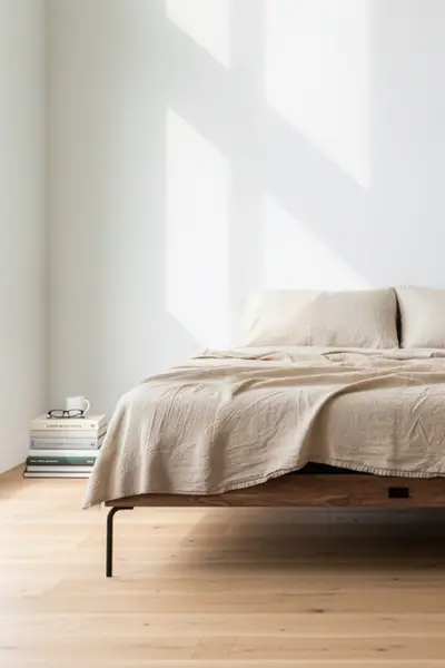 Minimalist bedroom with a low wooden bed in beige linens, a stack of books and glasses beside it, sunlight casting geometric shadows.